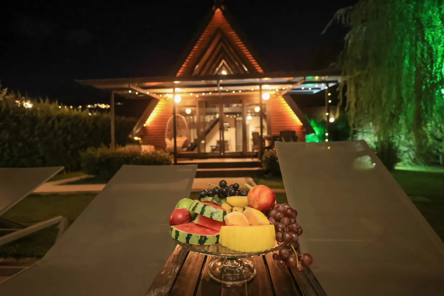 Gece ışıkları yanan A-frame bungalov, önde şezlonglar ve meyve tabağı