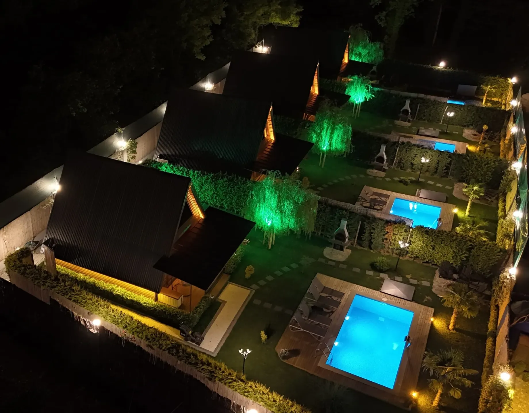 Gece yandan A-frame bungalovlar: ışıklı saçaklar, yeşil aydınlatılmış ağaçlar ve mavi havuzlar