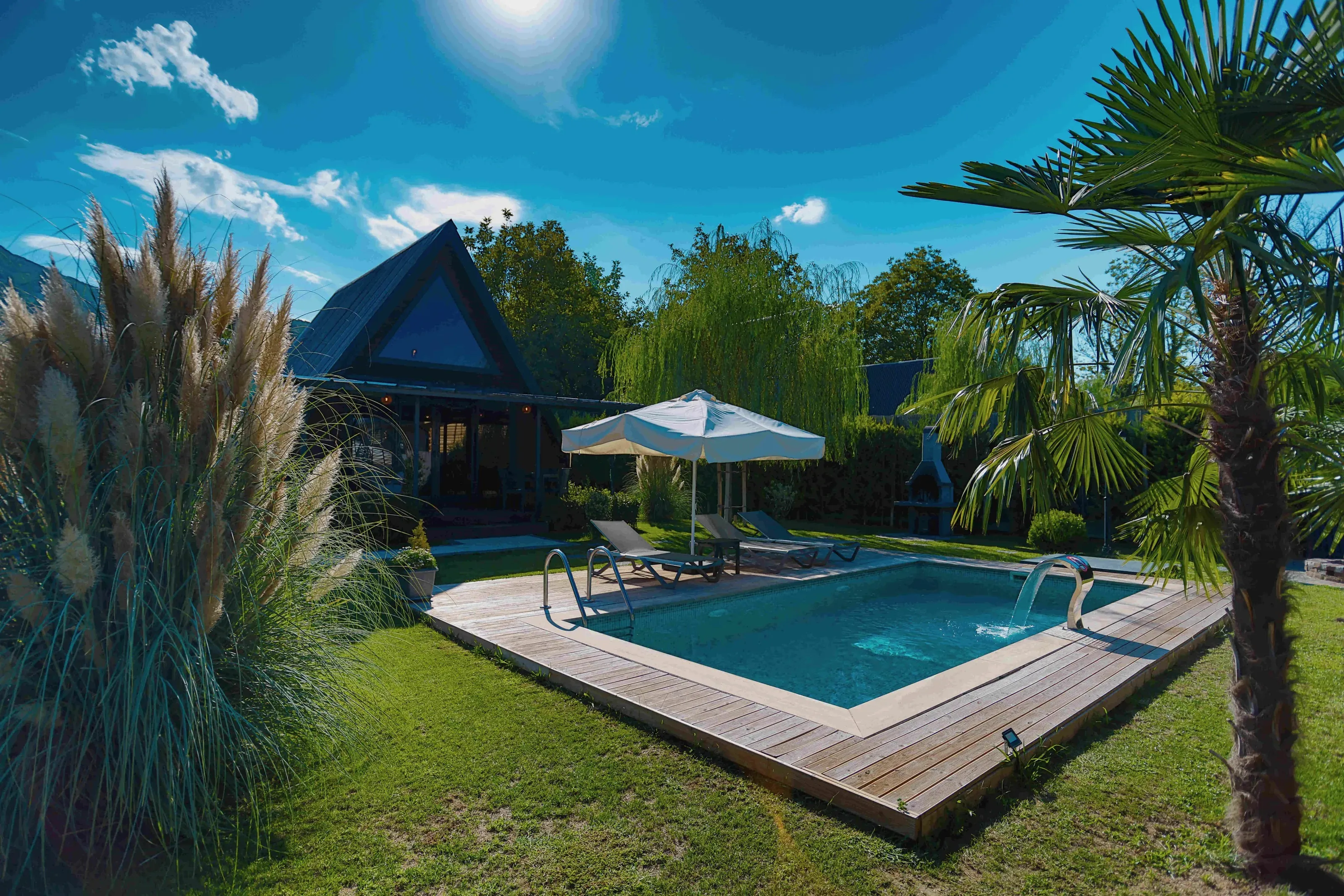 Siyah A-frame bungalov, önünde ahşap güverteli havuz, pampas otları ve palmiye, açık gök